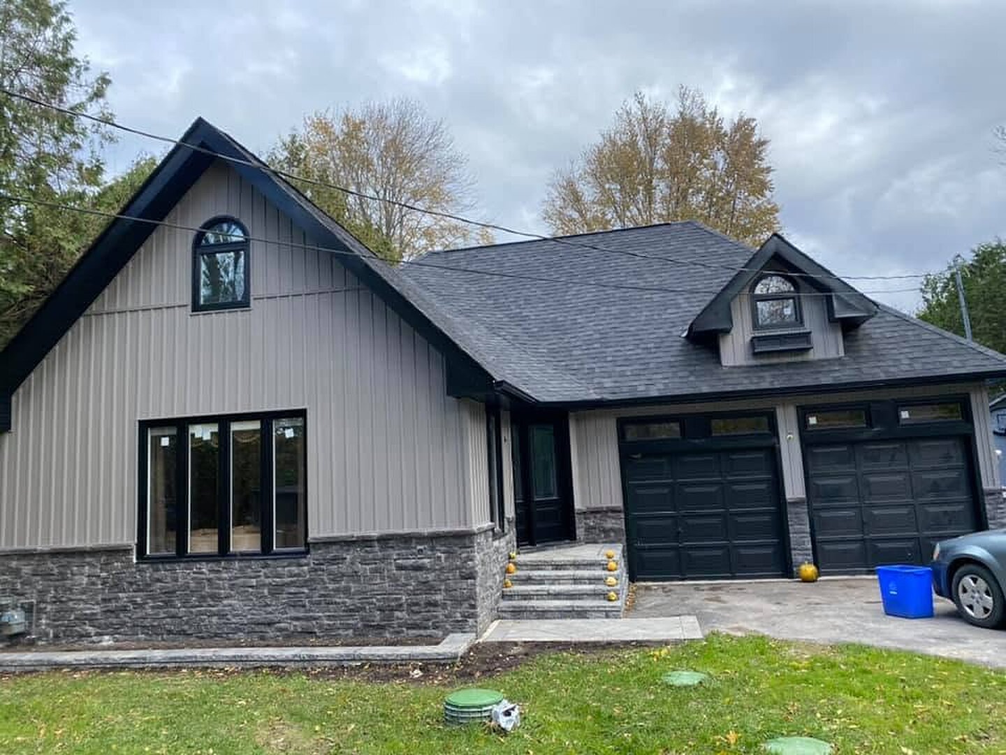 Finished home exterior with taupe board-and-batten siding, black windows, black garage doors, and a stone base.